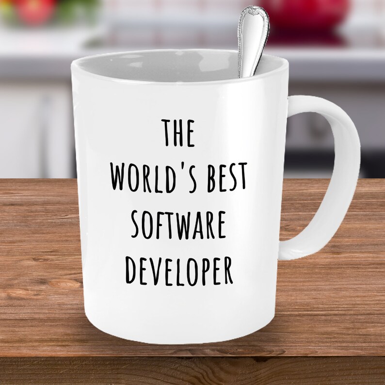 May include: White ceramic mug with the text "THE WORLD'S BEST SOFTWARE DEVELOPER" in black. A silver spoon is inside the mug. The mug is on a wooden surface.