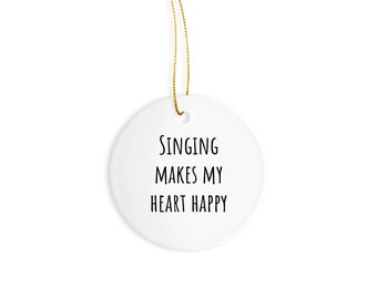 Może przedstawiać: Biała ozdoba ceramiczna ze złotym sznurkiem do zawieszenia. Ozdoba ma czarny napis "Singing makes my heart happy."