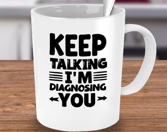 May include: White ceramic mug with the text "KEEP TALKING I'M DIAGNOSING YOU" in black. A silver spoon is inside the mug. The mug is on a wooden surface.