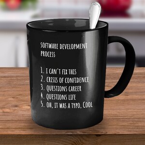 May include: Black coffee mug with white text that reads "SOFTWARE DEVELOPMENT PROCESS" and a numbered list: "1. I CAN'T FIX THIS 2. CRISIS OF CONFIDENCE 3. QUESTIONS CAREER 4. QUESTIONS LIFE 5. OH, IT WAS A TYPO, COOL"