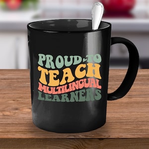 May include: Black coffee mug with a white handle. The mug has the text "Proud to Teach Multilingual Learners" in a retro font with colorful letters.
