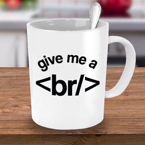 Op de afbeelding: Witte keramische koffiemok met een zwarte afbeelding waarop "give me a <br/>" staat, gedrukt op de voorkant.