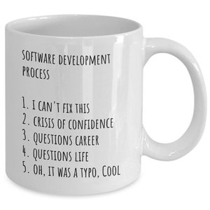 May include: White ceramic mug with black text that reads "SOFTWARE DEVELOPMENT PROCESS" and a numbered list: "1. I CAN'T FIX THIS, 2. CRISIS OF CONFIDENCE, 3. QUESTIONS CAREER, 4. QUESTIONS LIFE, 5. OH, IT WAS A TYPO, COOL"