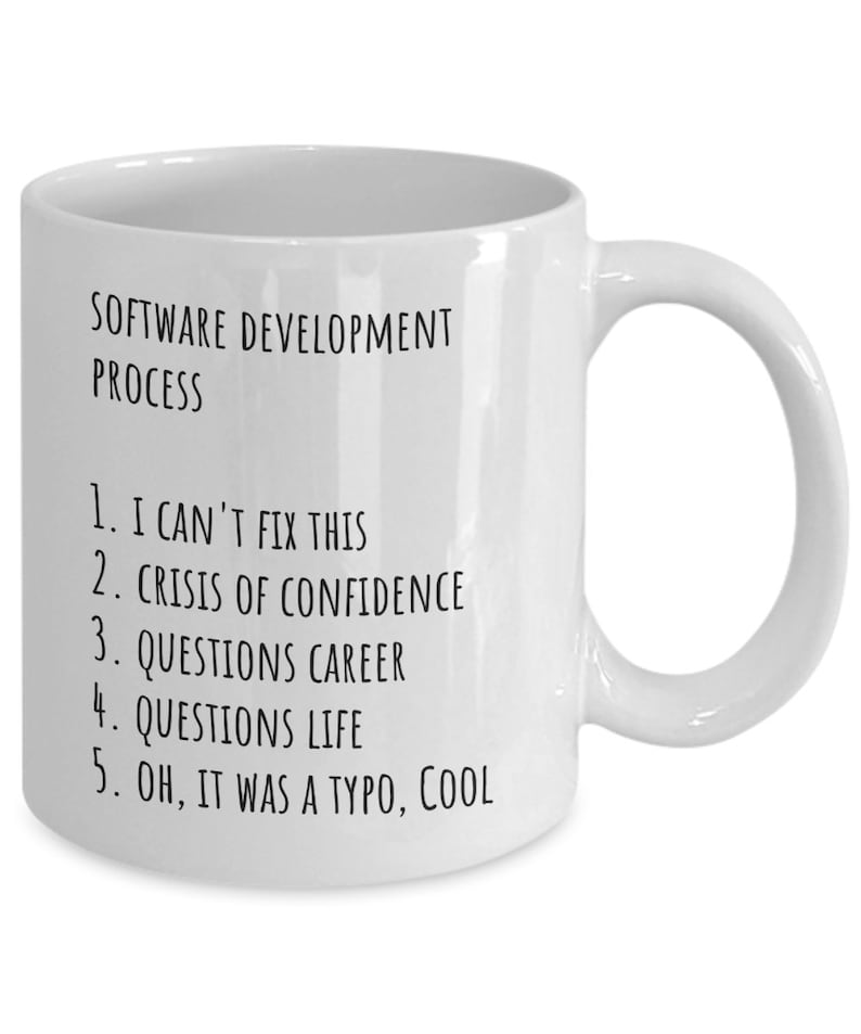 May include: White ceramic mug with black text that reads "SOFTWARE DEVELOPMENT PROCESS" and a numbered list: "1. I CAN'T FIX THIS, 2. CRISIS OF CONFIDENCE, 3. QUESTIONS CAREER, 4. QUESTIONS LIFE, 5. OH, IT WAS A TYPO, COOL"