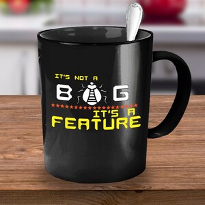 Puede incluir: Taza de café negra con el texto "IT'S NOT A BUG IT'S A FEATURE" en blanco y amarillo. Una cuchara plateada descansa dentro de la taza. La taza está sobre una superficie de madera.