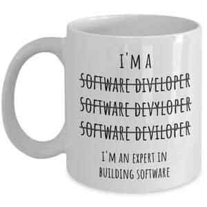 Op de afbeelding: Witte keramische mok met een gebogen handvat. De mok heeft de tekst "I'M A SOFTWARE DIVELOPER" doorgestreept, gevolgd door "SOFTWARE DEVYLOPER" doorgestreept, dan "SOFTWARE DEVILOPER" doorgestreept. Daaronder staat de tekst "I'M AN EXPERT IN BUILDING SOFTWARE."