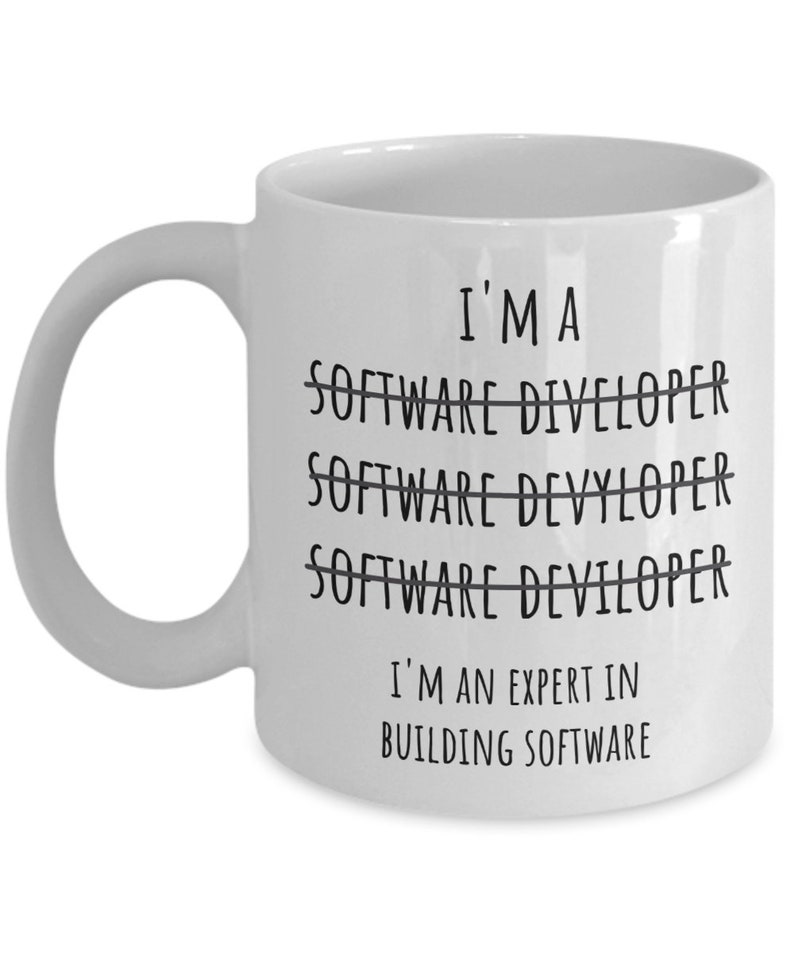 Op de afbeelding: Witte keramische mok met een gebogen handvat. De mok heeft de tekst "I'M A SOFTWARE DIVELOPER" doorgestreept, gevolgd door "SOFTWARE DEVYLOPER" doorgestreept, dan "SOFTWARE DEVILOPER" doorgestreept. Daaronder staat de tekst "I'M AN EXPERT IN BUILDING SOFTWARE."