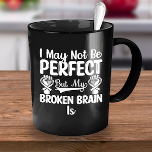 May include: Black coffee mug with white text that reads "I May Not Be Perfect But My Broken Brain Is" with a white ribbon and fist graphic on each side.