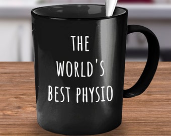 Könnte beinhalten: Schwarze Keramik-Tasse mit dem weißen Text "THE WORLD'S BEST PHYSIO". Ein silberner Löffel liegt in der Tasse. Die Tasse steht auf einer Holzoberfläche.