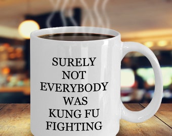 Puede incluir: Taza de café de cerámica blanca llena de líquido oscuro. La taza tiene un asa curva y el texto "SURELY NOT EVERYBODY WAS KUNG FU FIGHTING" en negro. El fondo está borroso.
