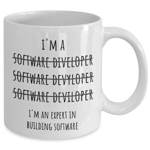 Op de afbeelding: Witte keramische mok met de zwarte tekst "I'M A SOFTWARE DIVELOPER SOFTWARE DEVYLOPER SOFTWARE DEVILOPER I'M AN EXPERT IN BUILDING SOFTWARE". De mok heeft een gebogen oor en is ontworpen voor warme dranken.