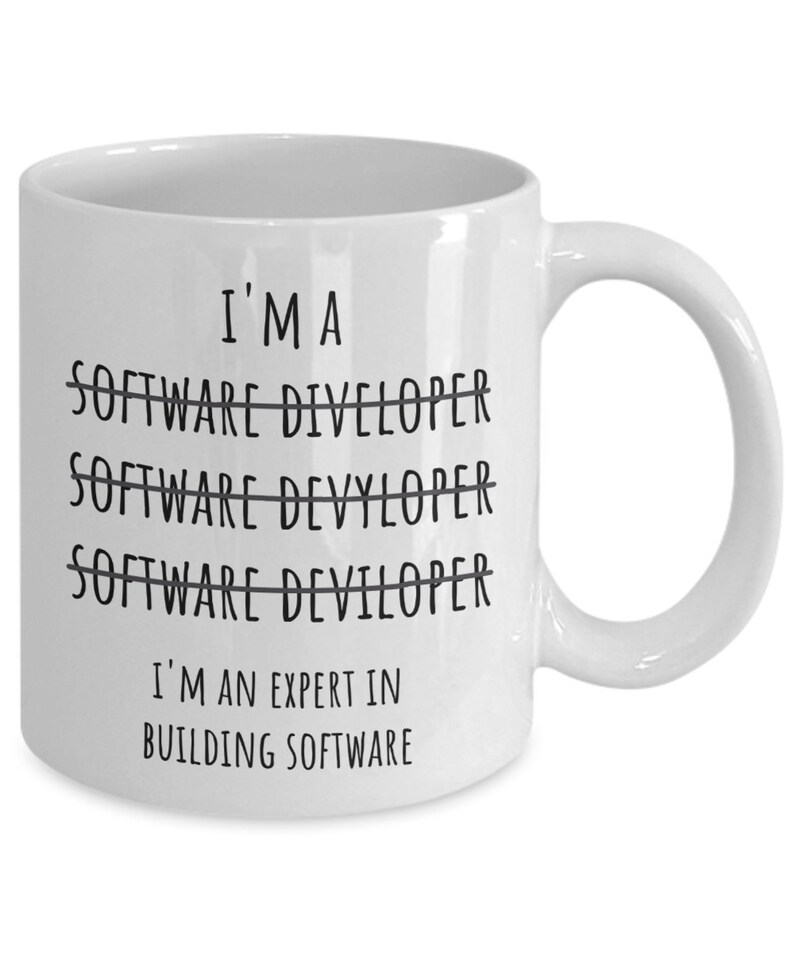 Op de afbeelding: Witte keramische mok met de zwarte tekst "I'M A SOFTWARE DIVELOPER SOFTWARE DEVYLOPER SOFTWARE DEVILOPER I'M AN EXPERT IN BUILDING SOFTWARE". De mok heeft een gebogen oor en is ontworpen voor warme dranken.