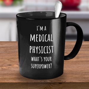 Puede incluir: Taza de café negra con texto blanco que dice "Soy un físico médico. ¿Cuál es tu superpoder?"