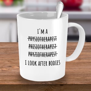 Puede incluir: Taza de cerámica blanca con asa curva y cuchara plateada en su interior. La taza tiene texto negro que dice "I'M A PHYSIOTHERAPIST" con la palabra "PHYSIOTHERAPIST" tachada tres veces, y "I LOOK AFTER BODIES".