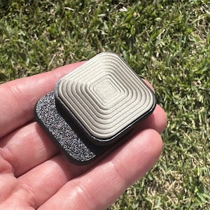 Op de afbeelding: Een zilverkleurig vierkant object met concentrische cirkels op het bovenoppervlak. Het object is bevestigd aan een zwarte vierkante basis met een getextureerd oppervlak.