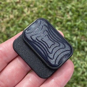 May include: A black phone grip with a textured, wavy design. The grip has a sparkly, iridescent finish.