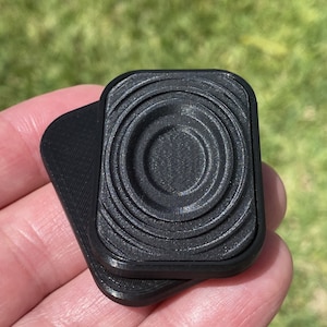 May include: Two black plastic square-shaped objects with a concentric circle design on the top. The objects are stacked on top of each other.