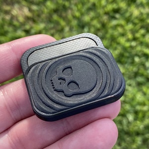 May include: A black square-shaped phone grip with a textured surface and a skull design. The grip has a smooth, silver-colored surface on the back.