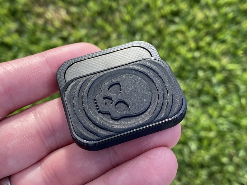 May include: A black square-shaped phone grip with a textured surface and a skull design. The grip has a smooth, silver-colored surface on the back.