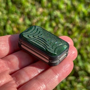 Könnte beinhalten: Kleine, rechteckige, dunkelgrüne Box mit einem strukturierten, wellenartigen Design auf der Oberseite. Die Box hat silberfarbene Metallseiten und wird in der Hand einer Person vor einem verschwommenen grünen Hintergrund gehalten.