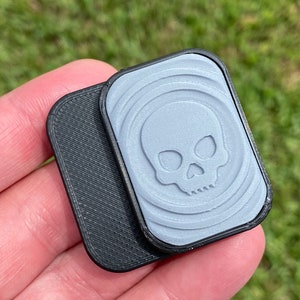 May include: Two small, rectangular, 3D printed objects. One is black with a textured surface. The other is light gray with a skull design and a textured surface.