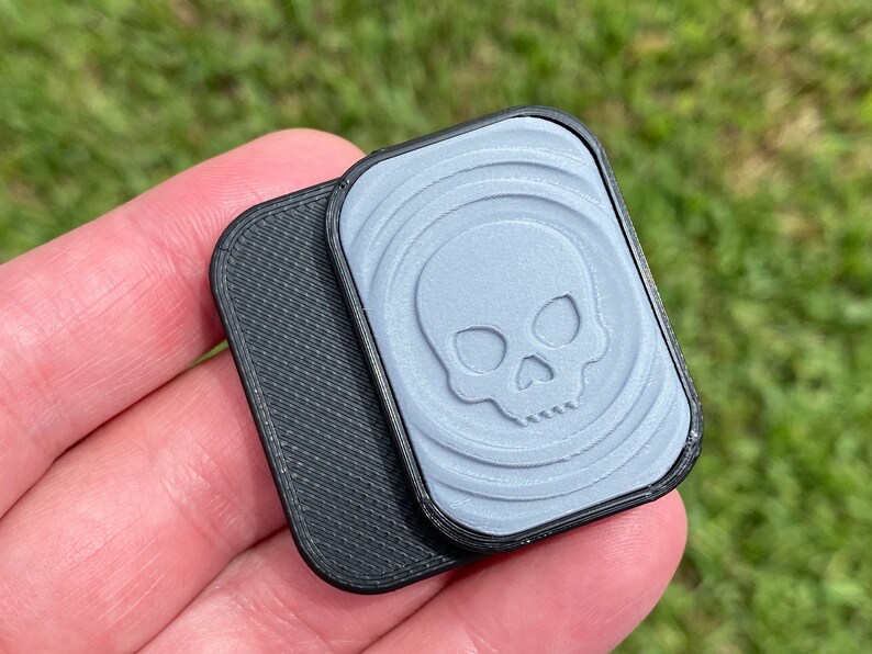 May include: Two small, rectangular, 3D printed objects. One is black with a textured surface. The other is light gray with a skull design and a textured surface.