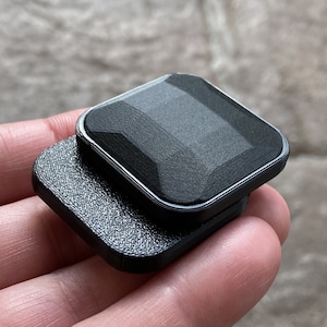 Puede incluir: Un agarre para teléfono de color negro con forma cuadrada y superficie texturizada. El agarre tiene un diseño en forma de diamante elevado en la parte superior.