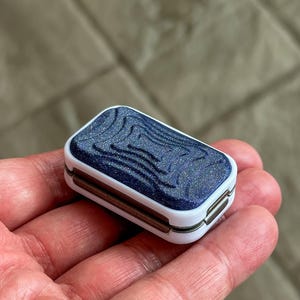 May include: A small rectangular container with a white frame and a dark blue, glittery, textured lid featuring concentric, wavy, topographical-style lines. The sides of the container are white with a metallic bronze-colored hinge mechanism visible.