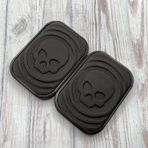 May include: Two black rectangular coasters with a skull design embossed in the center. The coasters have a textured surface.