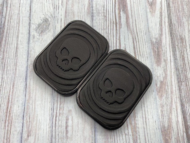 May include: Two black rectangular coasters with a skull design embossed in the center. The coasters have a textured surface.