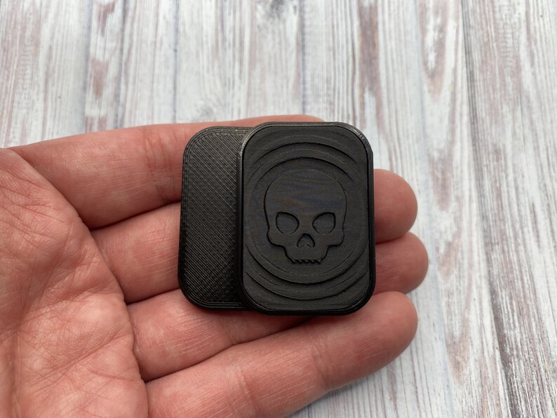 May include: A black wooden square with a skull design carved into the surface. The square is held in a hand.