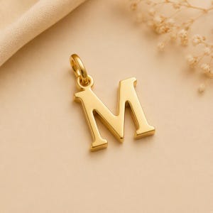 Könnte beinhalten: Ein polierter goldfarbener Metallanhänger in Form des Großbuchstabens M. Der Anhänger hat eine glatte, reflektierende Oberfläche und verfügt über einen kleinen runden Verbindungsring an der Oberseite zur Befestigung an einer Kette oder Schnur.
