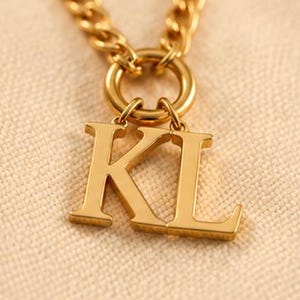 Puede incluir: Un collar de tono dorado con una cadena de metal pulido y un anillo conector circular. Dos dijes de letras mayúsculas, K y L, cuelgan del anillo. Las letras tienen una superficie lisa y reflectante con un estilo de fuente serif clásico.