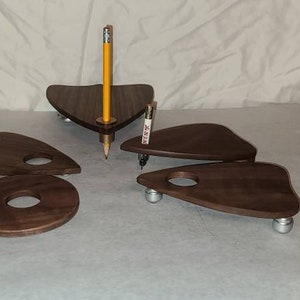 Puede incluir: Cuatro posavasos de madera con forma de triángulo y bordes redondeados. Los posavasos son de madera oscura y tienen un acabado liso. Los posavasos están dispuestos en un patrón sobre una superficie blanca. Un lápiz amarillo está de pie en el centro de los posavasos.
