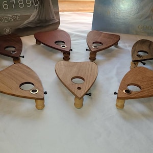 May include: Six wooden planchette boards for a Ouija board game. The planchette boards are triangular shaped and have a hole in the centre. The boards are made of different types of wood, including light and dark wood. The boards are arranged in a row on a white surface.