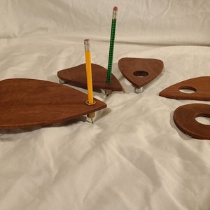 May include: Wooden planchette set for a Ouija board game. The set includes four planchette boards, two of which are shaped like a teardrop, one is round, and one is heart-shaped. The planchette boards are made of dark brown wood and have a hole in the center. Two yellow pencils are placed on the planchette boards.