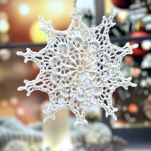 Könnte beinhalten: Ein weißer, gehäkelter Schneeflocken-Anhänger mit einem filigranen, komplizierten Design. Die Schneeflocke hängt an einer weißen Schnur.