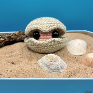 May include: A crocheted clam with black eyes and a pink interior, sitting on a bed of sand with seashells.