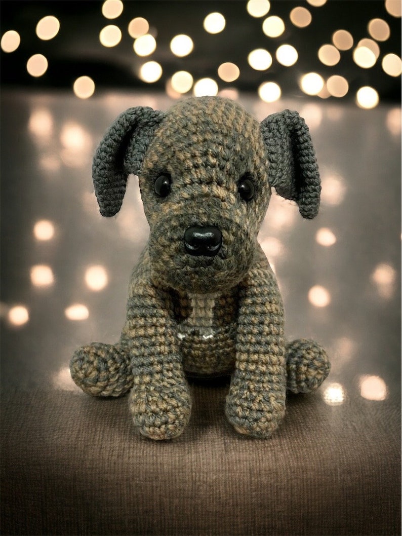 May include: A crocheted stuffed dog toy with brown and gray fur, sitting on a brown fabric surface. The dog has black eyes and a black nose.