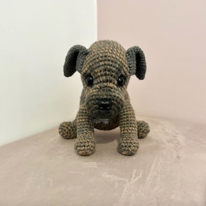 May include: A crocheted brown and gray puppy dog toy with black eyes and a black nose. The toy is sitting on a light brown surface.