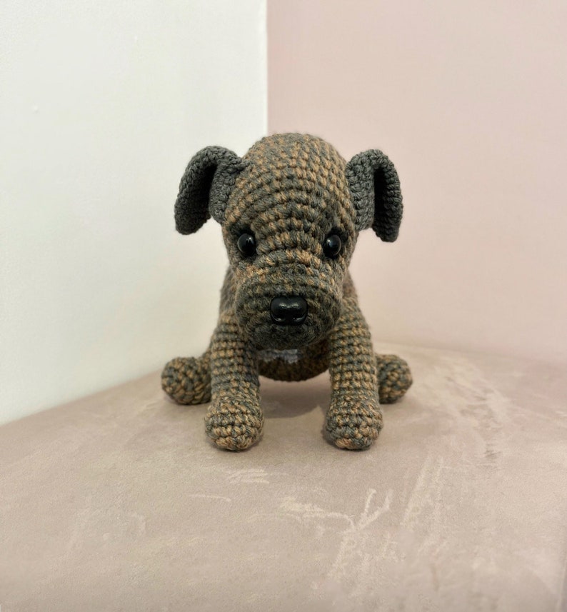 May include: A crocheted brown and gray puppy dog toy with black eyes and a black nose. The toy is sitting on a light brown surface.