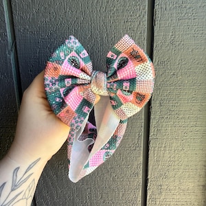 May include: A large, patterned hair bow featuring a patchwork design in shades of pink, green, and brown. The bow is held in a hand, showcasing its vibrant colors and intricate pattern. The bow's fabric has a textured appearance.