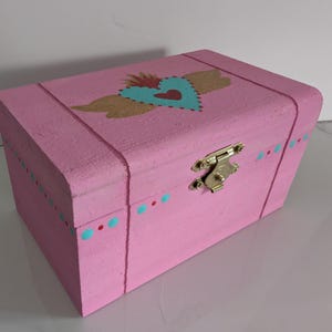 May include: A pink wooden box with a gold heart and wings design. The box has a gold latch and a turquoise and red polka dot pattern around the bottom.