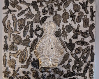 May include: A silver and black metal plaque covered in small charms. The charms are in the shape of hearts, crosses, angels, fish, leaves, and other religious symbols. The plaque features a large, silver, embossed image of the Virgin Mary in the center.