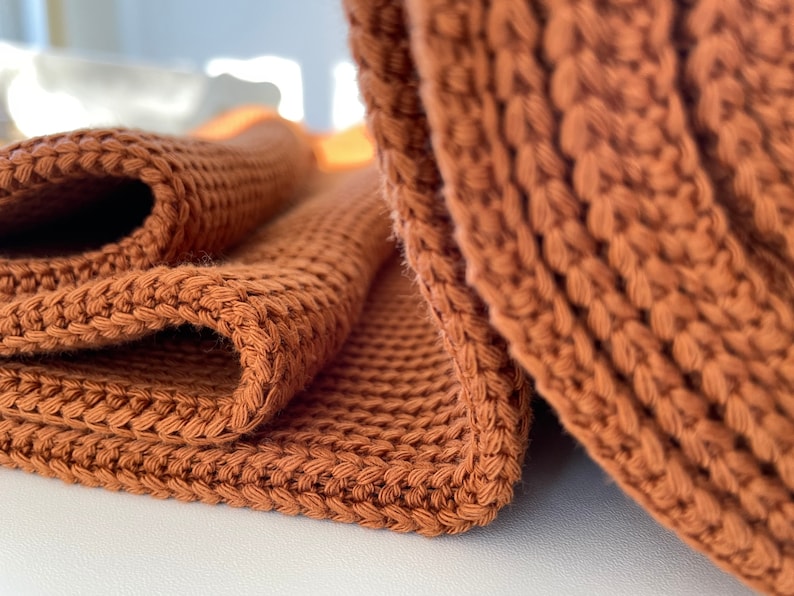 May include: A close-up of a brown knitted throw blanket folded in half. The blanket is made of a soft, textured yarn and has a subtle sheen.