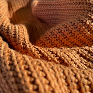 May include: Close-up of a brown knitted fabric with a textured pattern. The fabric is soft and warm, and the light catches the raised stitches.