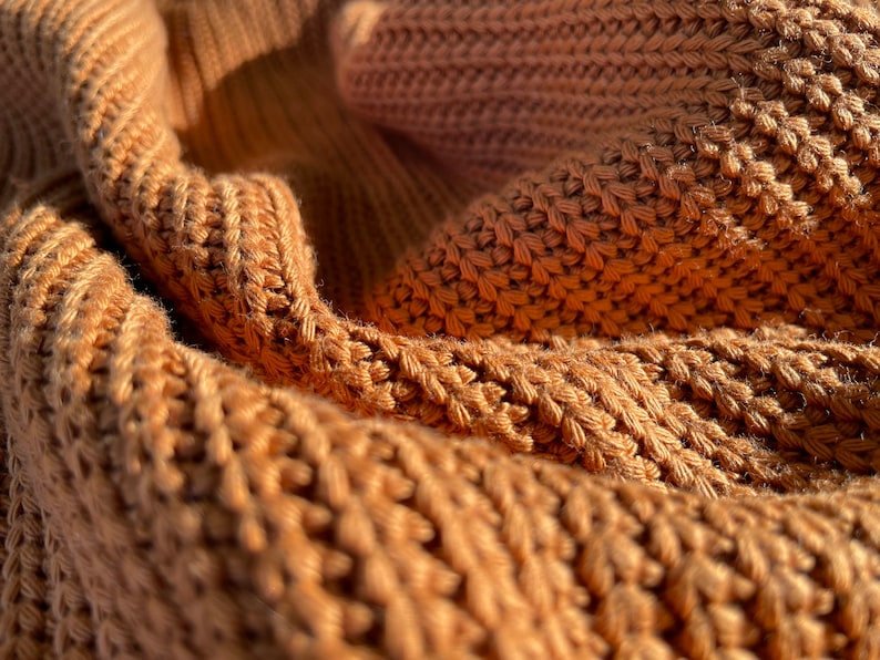 May include: Close-up of a brown knitted fabric with a textured pattern. The fabric is soft and warm, and the light catches the raised stitches.
