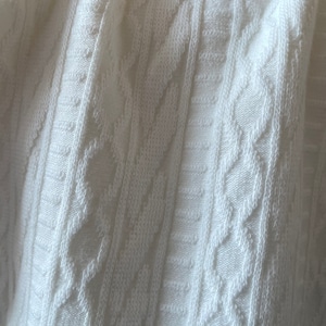 May include: Close-up of a white cable knit fabric. The texture features vertical and diagonal patterns, creating a textured surface. The fabric appears soft and is likely used for clothing or home decor items.