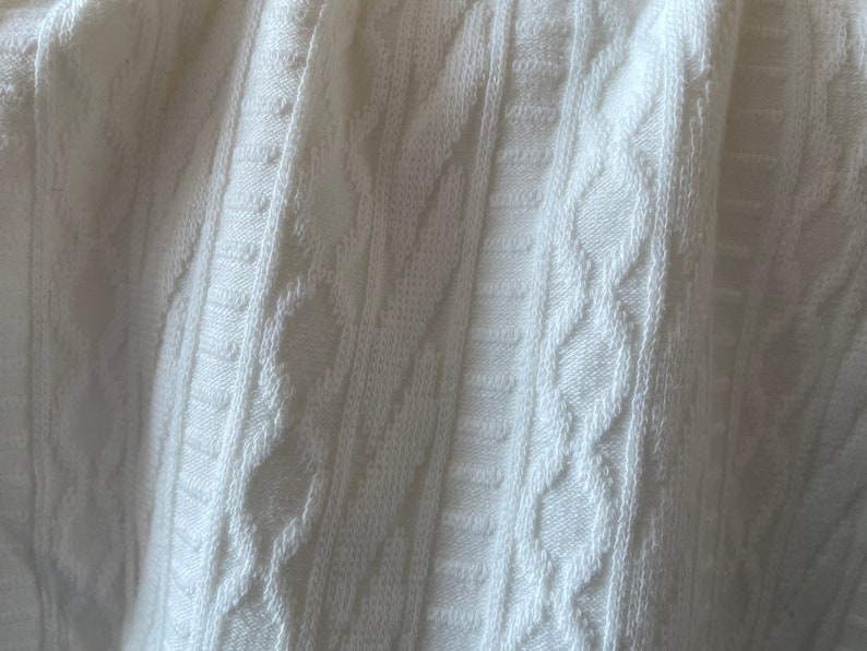 May include: Close-up of a white cable knit fabric. The texture features vertical and diagonal patterns, creating a textured surface. The fabric appears soft and is likely used for clothing or home decor items.