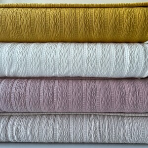 May include: Four folded blankets in a stack, each in a different colour. The top blanket is mustard yellow, followed by white, pale purple, and light grey. Each blanket features a textured cable-knit design.